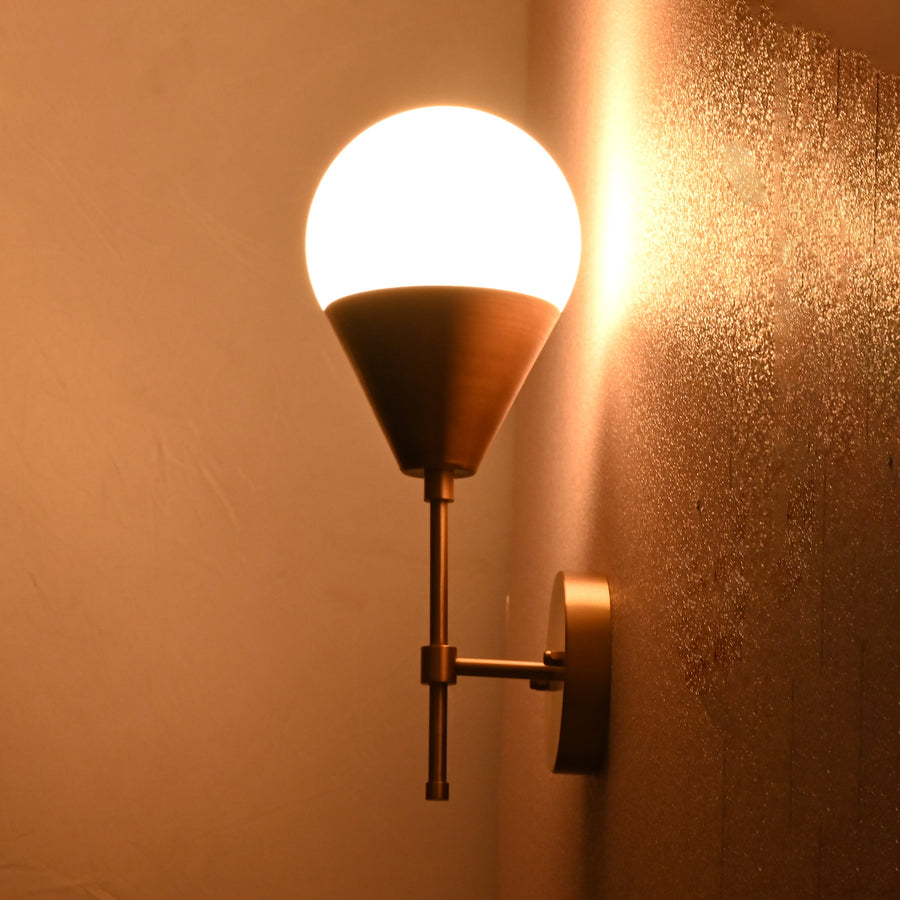 Modern brass wall sconce with globe shade and cone base in brushed brass finish, mounted on textured wall.