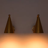 A pair of modern cone-shaped wall sconces in a brushed brass finish, mounted on a wall and casting a warm orange glow downwards.