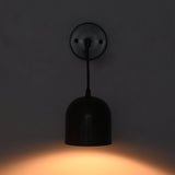 A modern black pendant wall light with a dome-shaped shade and circular wall plate, shown illuminated against a dark gray wall casting a warm orange glow.