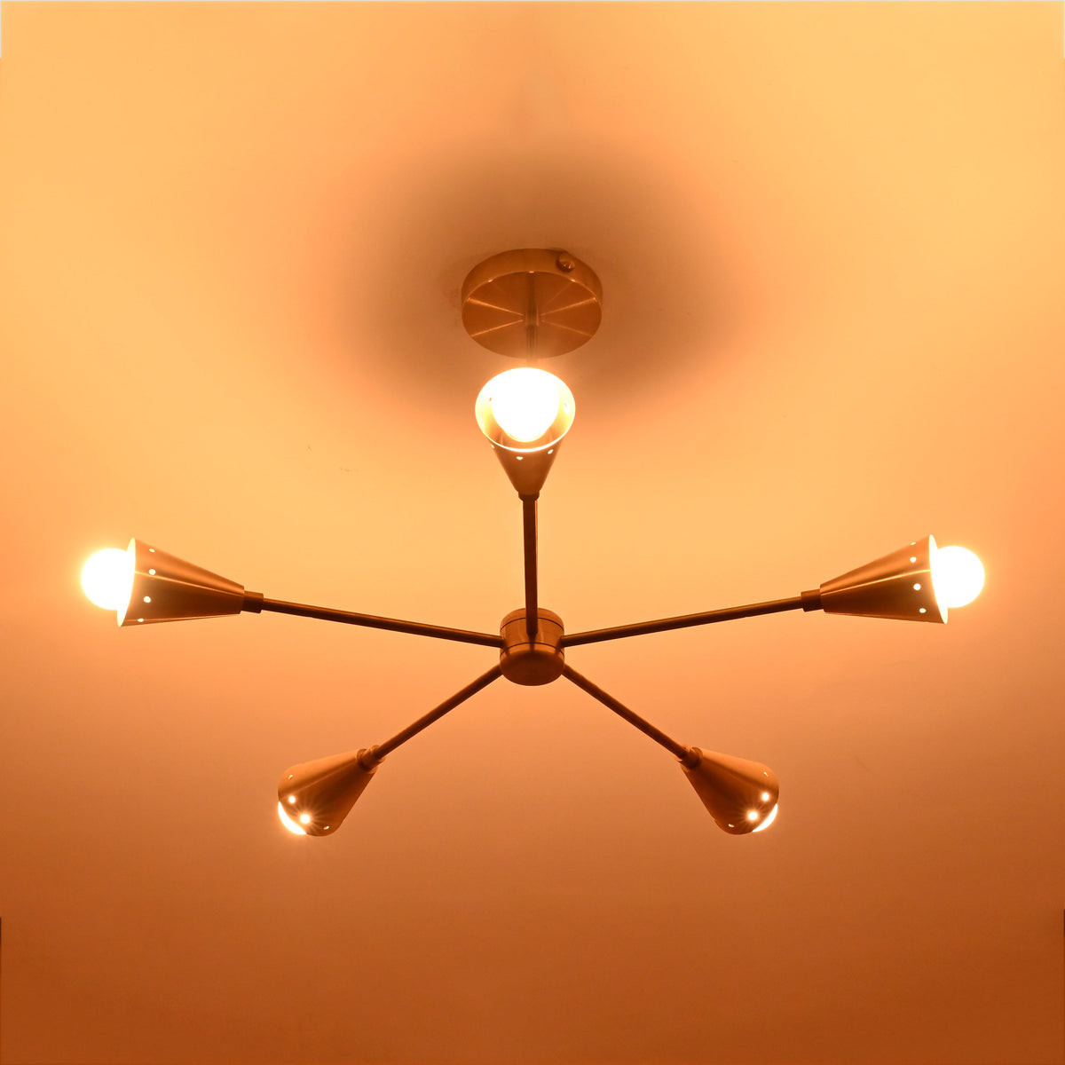 Lit view of handcrafted brass ceiling chandelier with five conical light arms, creating a golden modern glow in luxury interior spaces.