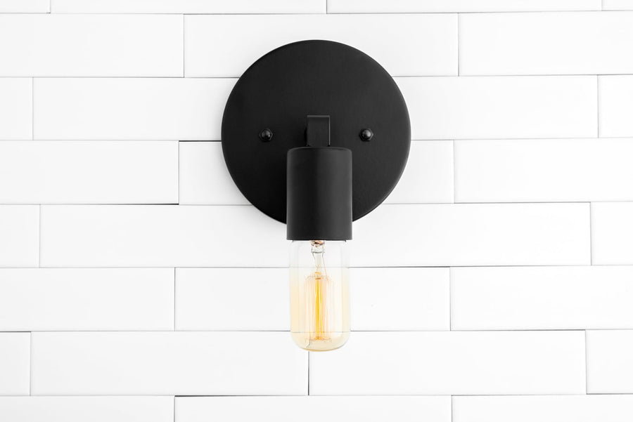Matte black wall sconce with round backplate and capsule Edison bulb on white subway tile wall