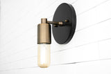 Modern wall sconce with matte black backplate and brushed brass cylinder socket holding capsule Edison bulb on white tile wall