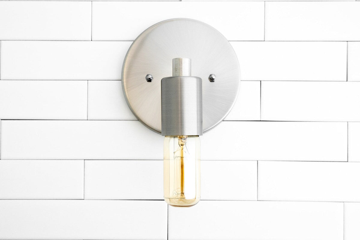 Brushed nickel wall sconce front view with round backplate and capsule Edison bulb on white tiled wall