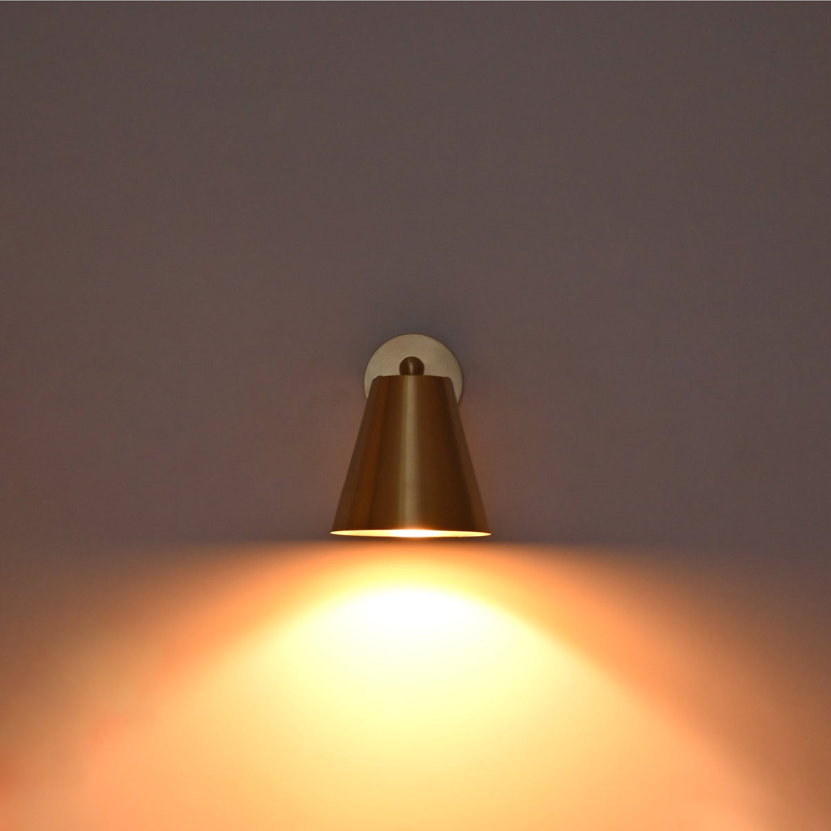 Front view of the Aurora satin brass tapered wall sconce illuminated with a warm downward glow creating soft ambient wall lighting.