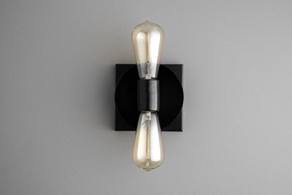 Contemporary wall sconce with black round disc on black square backplate and two vertical Edison-style bulbs, sleek minimalist industrial light.