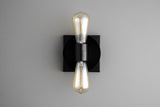 Contemporary wall sconce with black round disc on black square backplate and two vertical Edison-style bulbs, sleek minimalist industrial light.