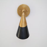 Front view of two-tone brass and black cone wall sconce with centered round brass backplate on white wall.