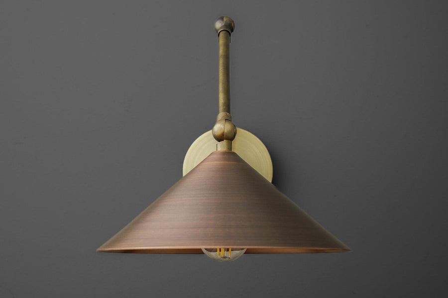 Adjustable brass swing arm wall sconce with cone shade and round backplate, modern industrial wall lamp on gray wall.