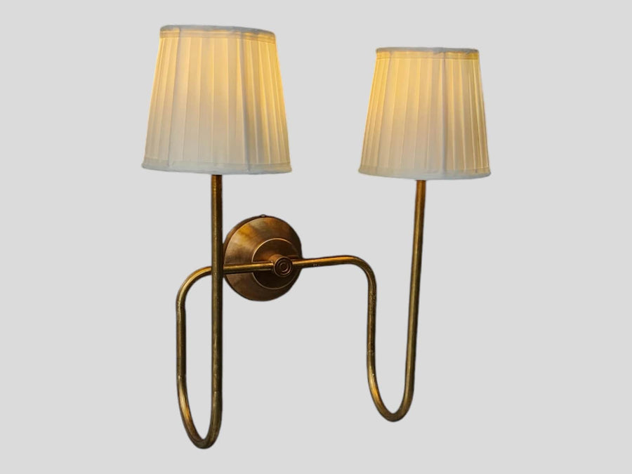 Side angled view of brass double arm wall sconce with pleated white shades, showing curved brass arms and warm ambient glow.
