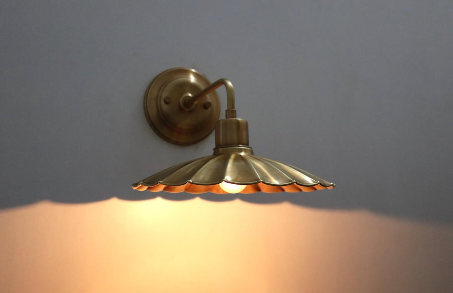 Lit brass wall sconce with scalloped disc shade casting a warm ambient glow on the wall below.