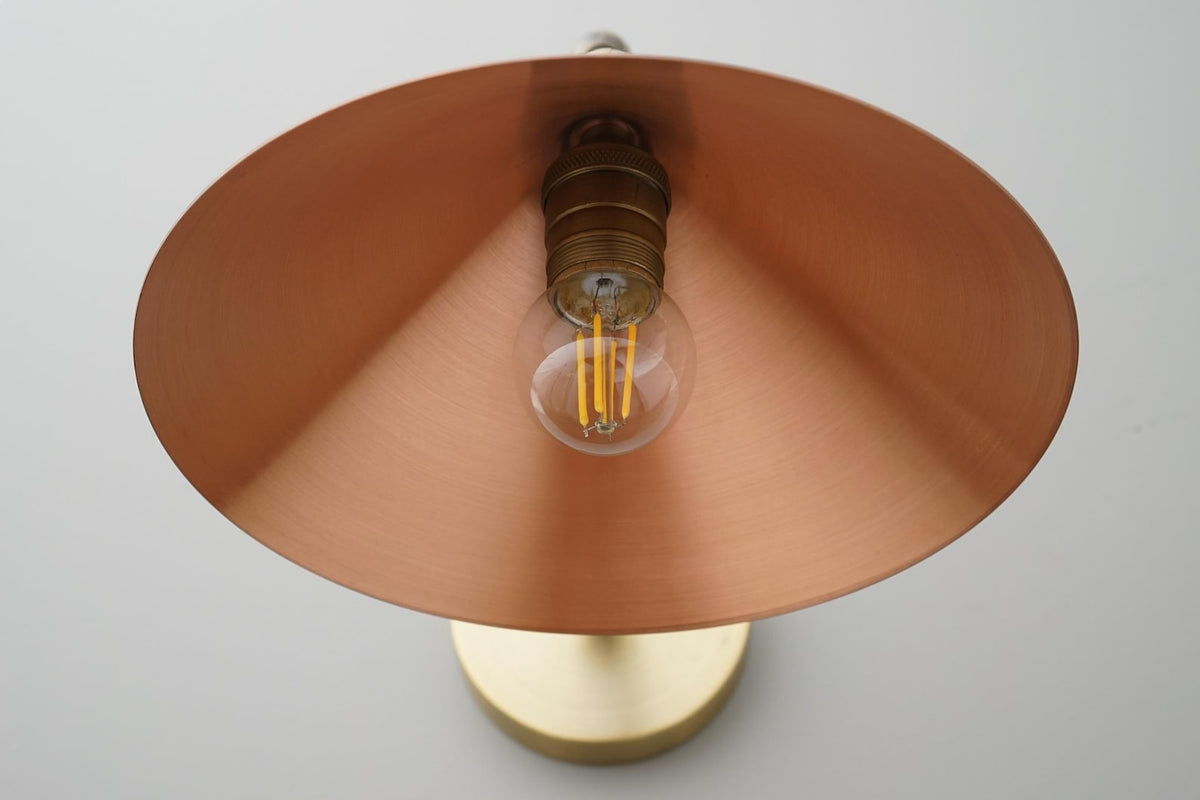 Close-up underside of brass cone shade showing warm copper-toned interior and exposed filament-style bulb, E26 wall sconce detail.