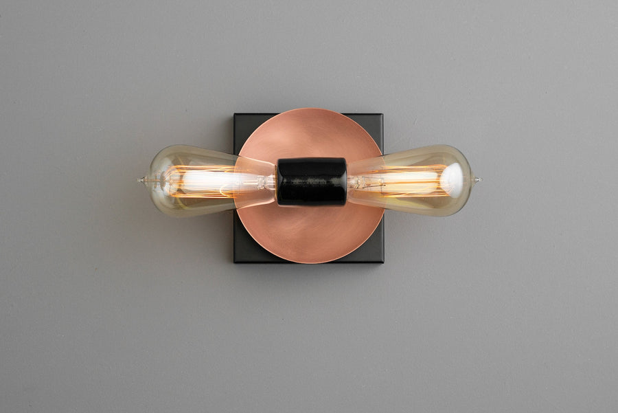 Modern wall sconce with copper disc backplate, black square base, and two vertical Edison-style bulbs, minimalist industrial lighting.