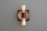 Modern wall sconce with copper disc backplate, black square base, and two vertical Edison-style bulbs, minimalist industrial lighting.