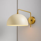 Brushed brass swing-arm dome wall sconce with cream metal shade mounted on a light gray wall.