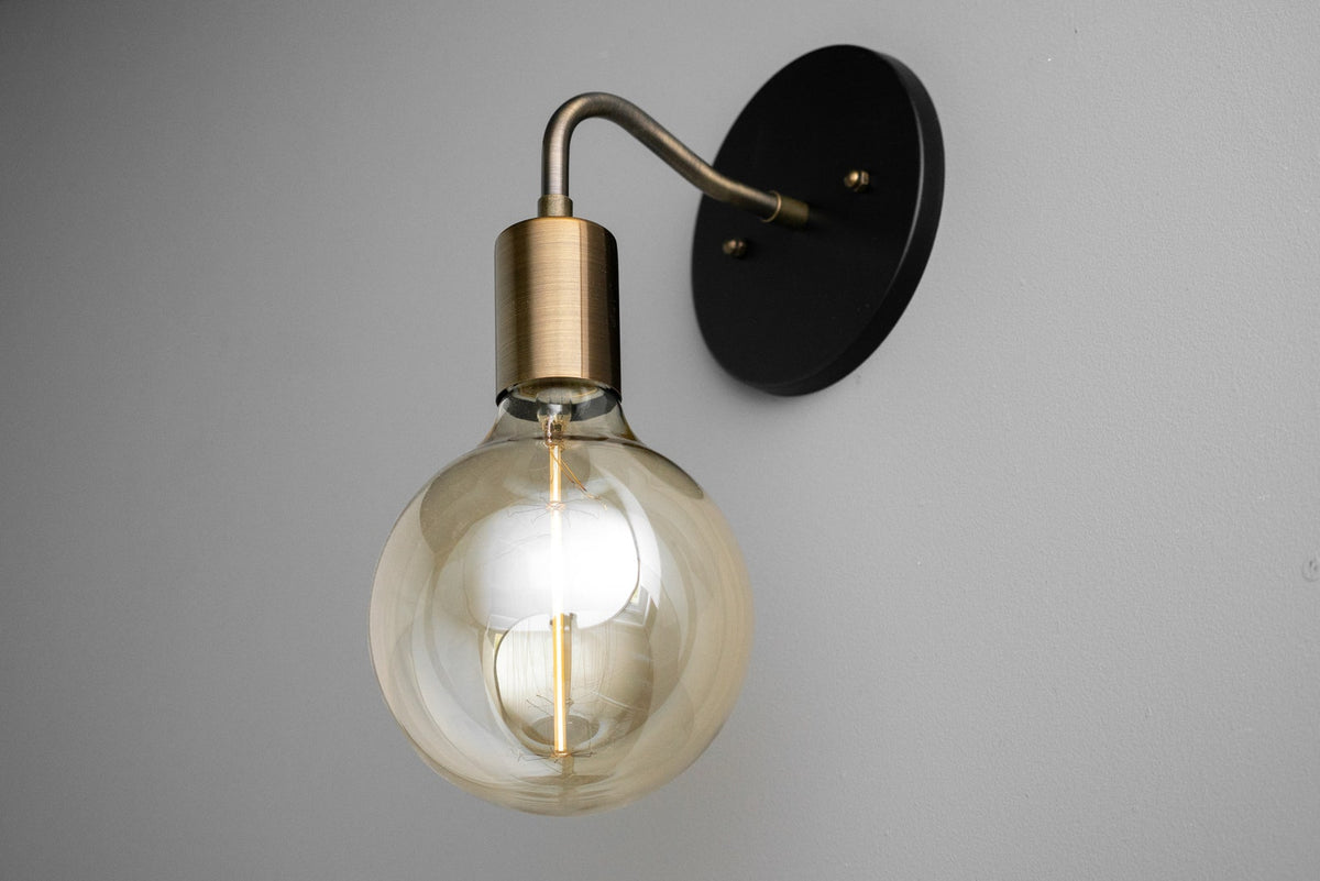 Modern brass and matte black wall sconce with smoked glass globe bulb on gray wall, minimalist industrial lighting fixture