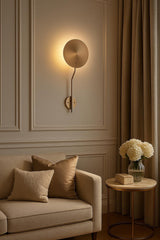 Brass disc wall sconce installed on paneled wall above sofa, creating soft ambient halo lighting in a warm neutral room.