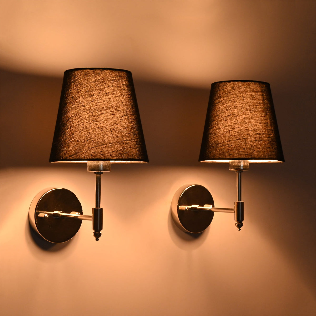 Side perspective of illuminated Milan polished chrome wall sconce with black tapered shade showcasing its warm ambient lighting effect.