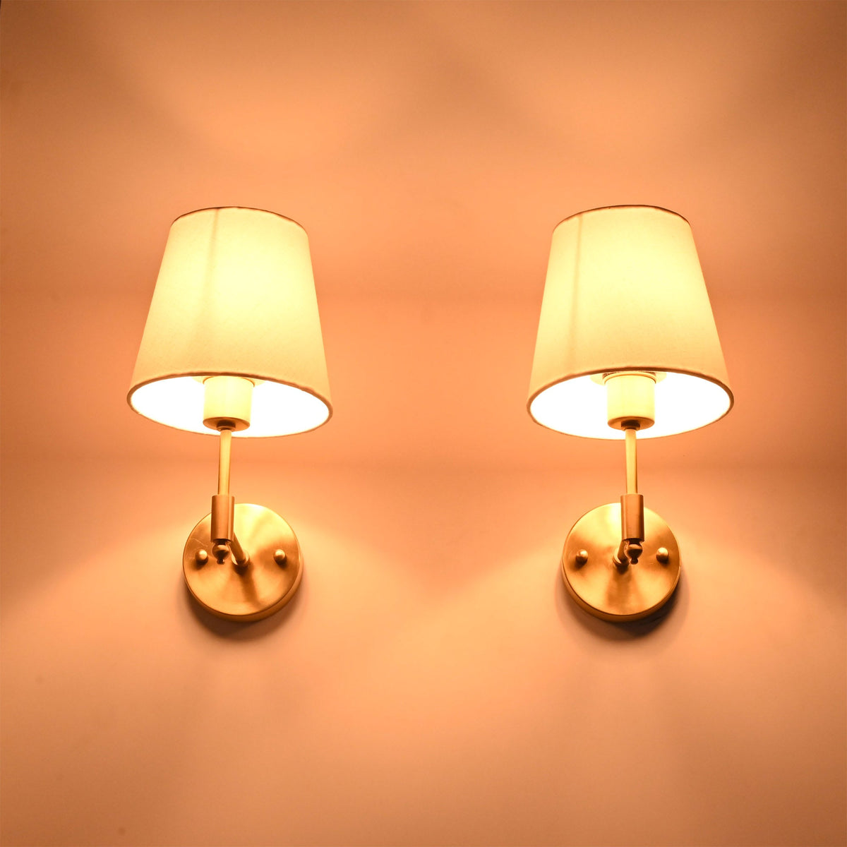 Illuminated Milan brushed brass wall sconces with white fabric shades casting a warm ambient glow against a neutral wall.