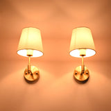 Illuminated Milan brushed brass wall sconces with white fabric shades casting a warm ambient glow against a neutral wall.