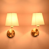 Perspective view of illuminated Milan brushed brass wall sconces with white fabric shades highlighting the handcrafted brass finish and soft lighting.