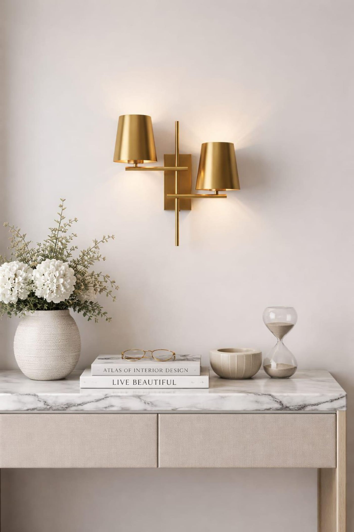 Modern 2-light brass wall sconce with dual tapered shades mounted above a marble-top console table with decor accents.