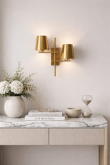 Modern 2-light brass wall sconce with dual tapered shades mounted above a marble-top console table with decor accents.