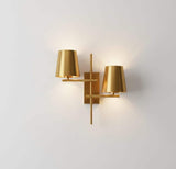 Front view of a geometric brass wall sconce with two tapered shades and warm ambient light on a white wall.