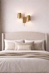 Brass 2-light wall sconce with twin tapered shades mounted above a bed in a soft neutral bedroom setting.