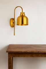 Single brushed brass wall sconce with curved arm and dome shade mounted on a white wall above a wooden table.
