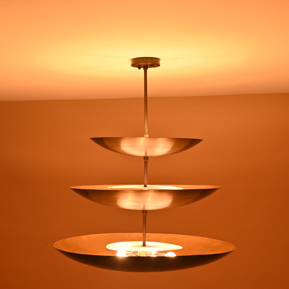 Full view of three-tier chandelier in blackened brass, glowing warmly with indirect upward illumination