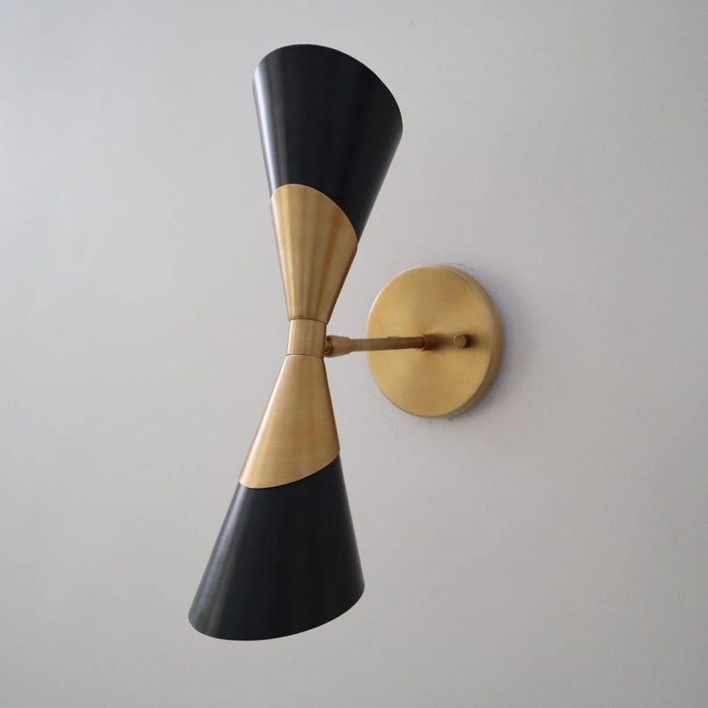 Angled right view of brass and black double cone wall sconce showing the round backplate and slim arm on a light wall.