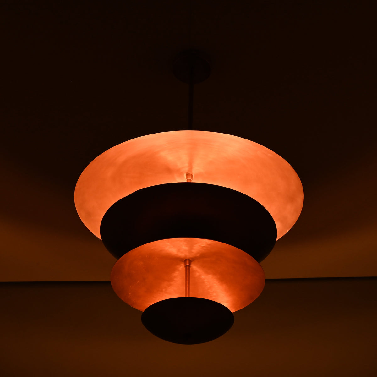 Night-shot of the brass bowl chandelier illuminated with warm indirect lighting across its stacked copper shades.
