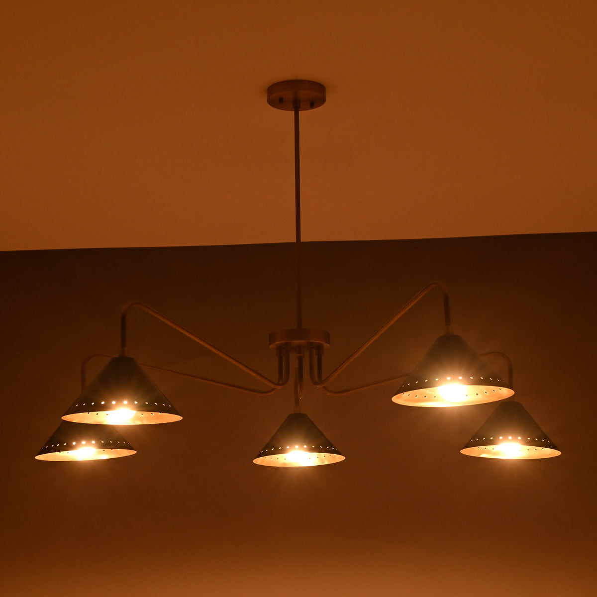 Evening ambience view of brass cone chandelier with five perforated black shades casting warm diffused light.