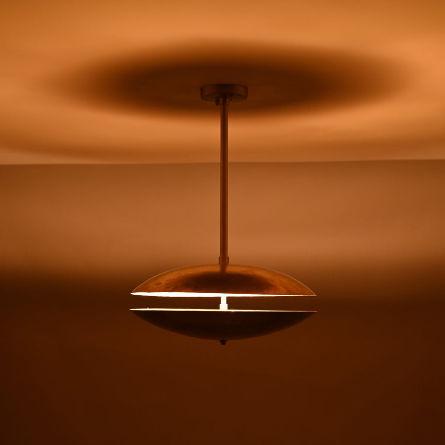 Handcrafted hammered brass disc pendant light with central rod suspension and warm indirect illumination, shown in a neutral interior setting.