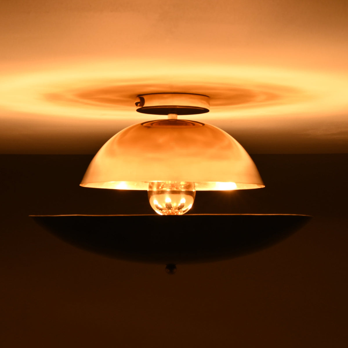 Exposed-bulb view of polished and blackened brass flushmount light, glowing warmly and casting soft diffused shadows on the ceiling.