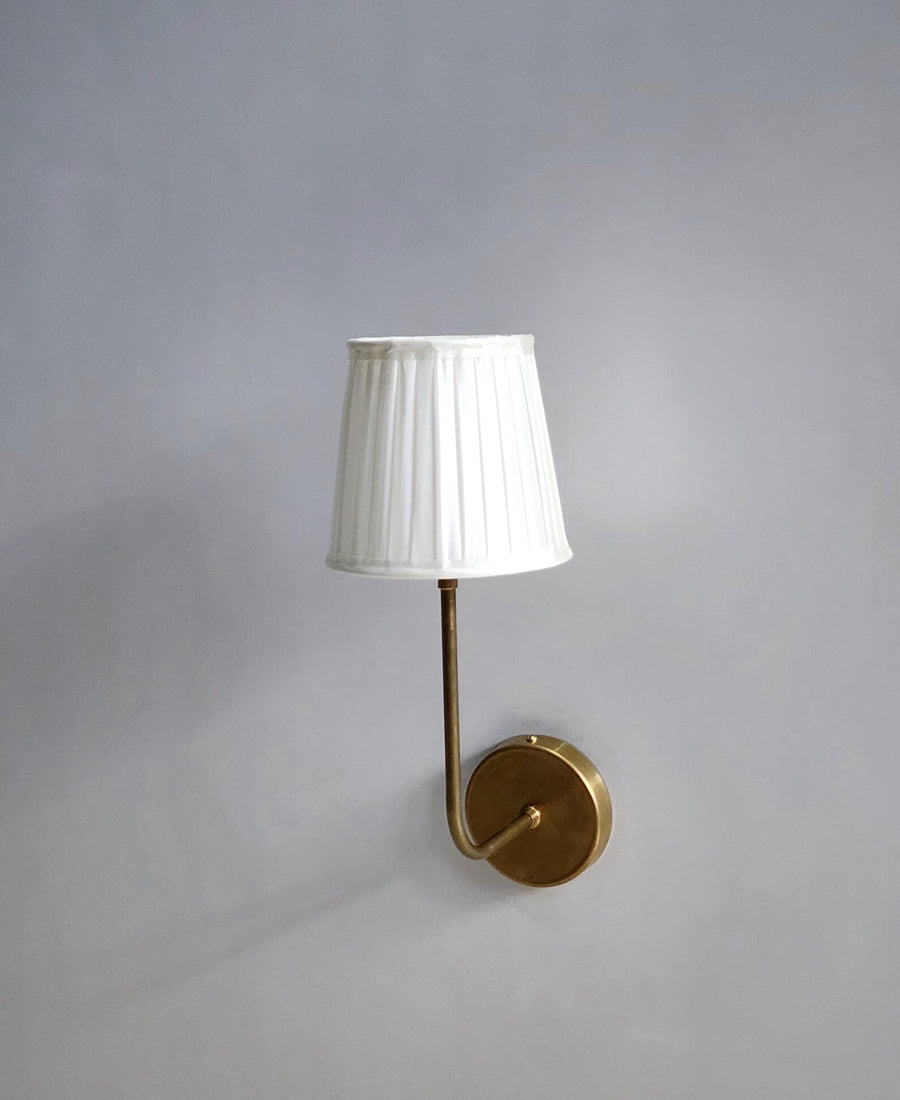 Handmade brass wall sconce by Brass Leaf Studio with pleated fabric shade glowing warmly against a wall.
