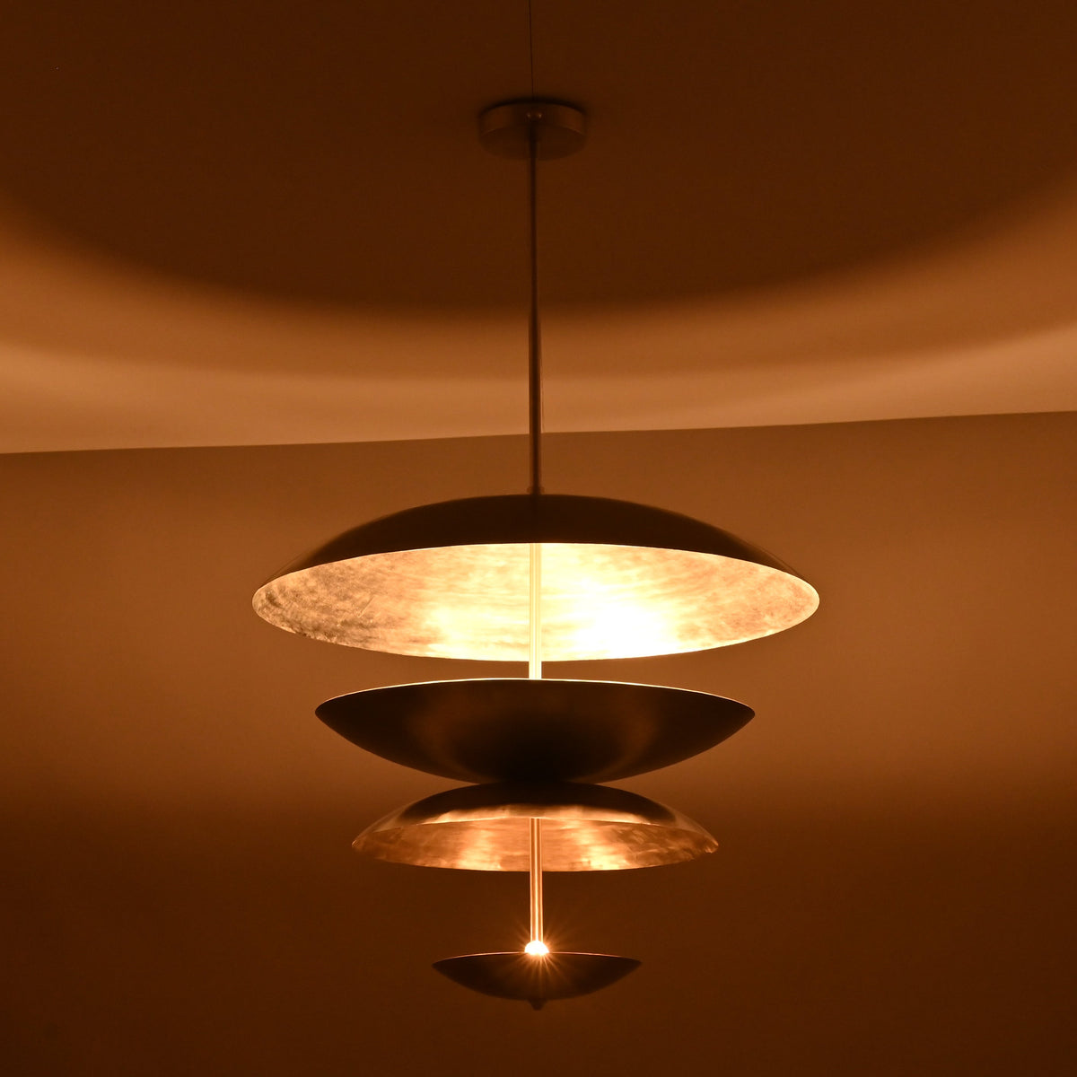 Night-view of a tiered metal disc pendant light displaying soft glowing reflections across layered metallic bowls.