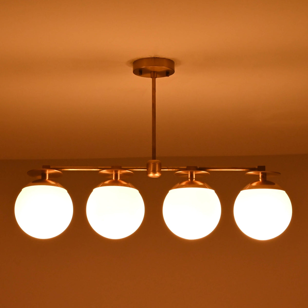 Front-facing view of brass linear chandelier with four opal glass globes in a low-light interior setting.
