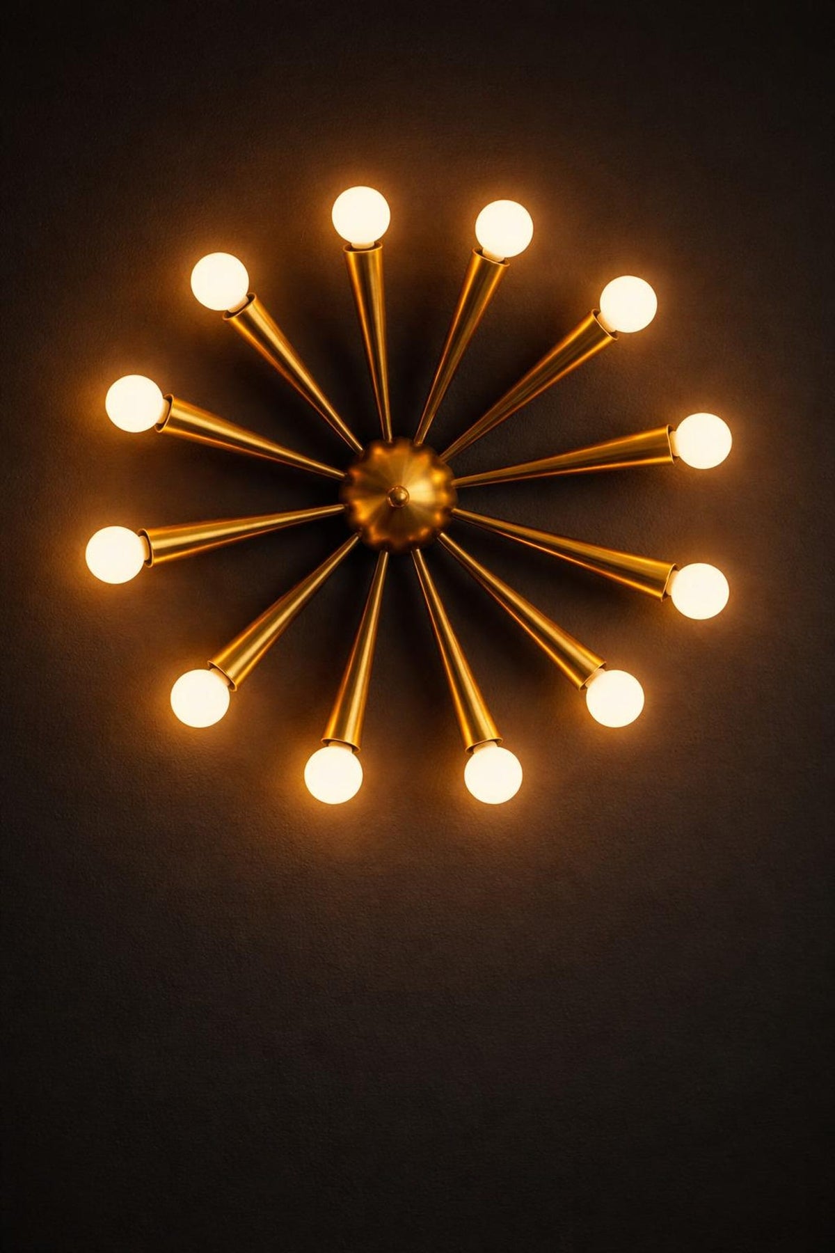 Brass starburst wall light glowing against a dark background, showcasing its warm ambient illumination and sculptural radial form.