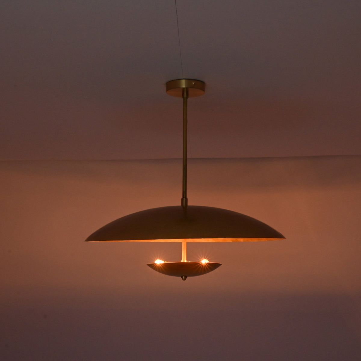 Brushed brass disc pendant illuminated in dark room creating soft indirect ambient lighting.