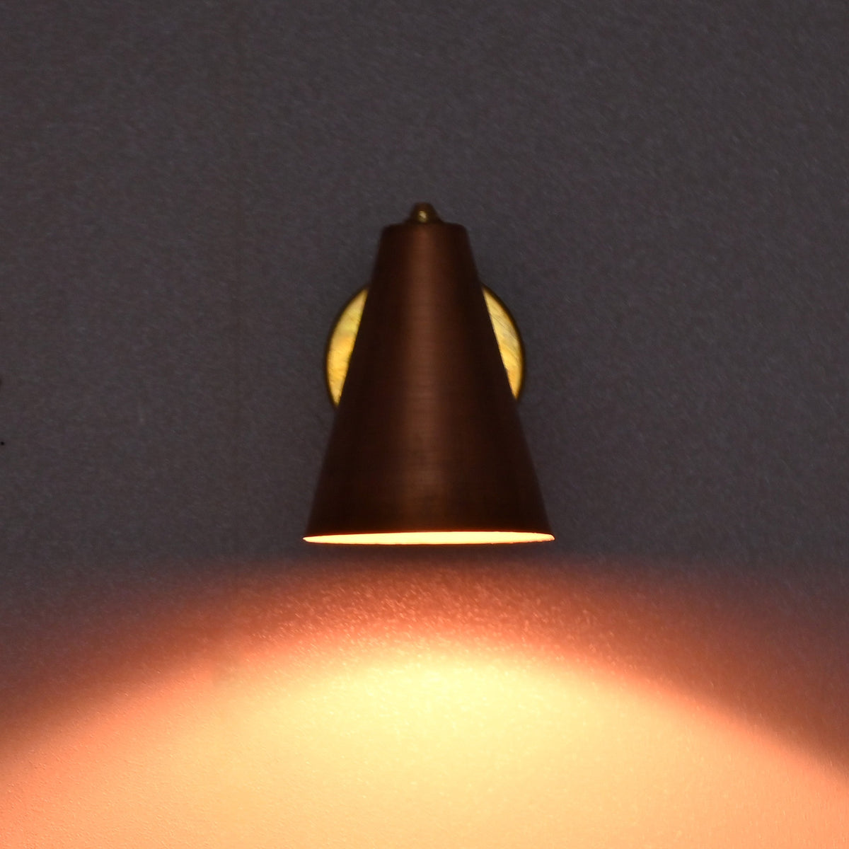 Brass and copper wall sconce lit in a dark setting, casting a warm ambient glow on the wall.