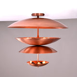 Front view of the lit brass flushmount chandelier showing warm indirect illumination across its stacked bowl-shaped copper shades.