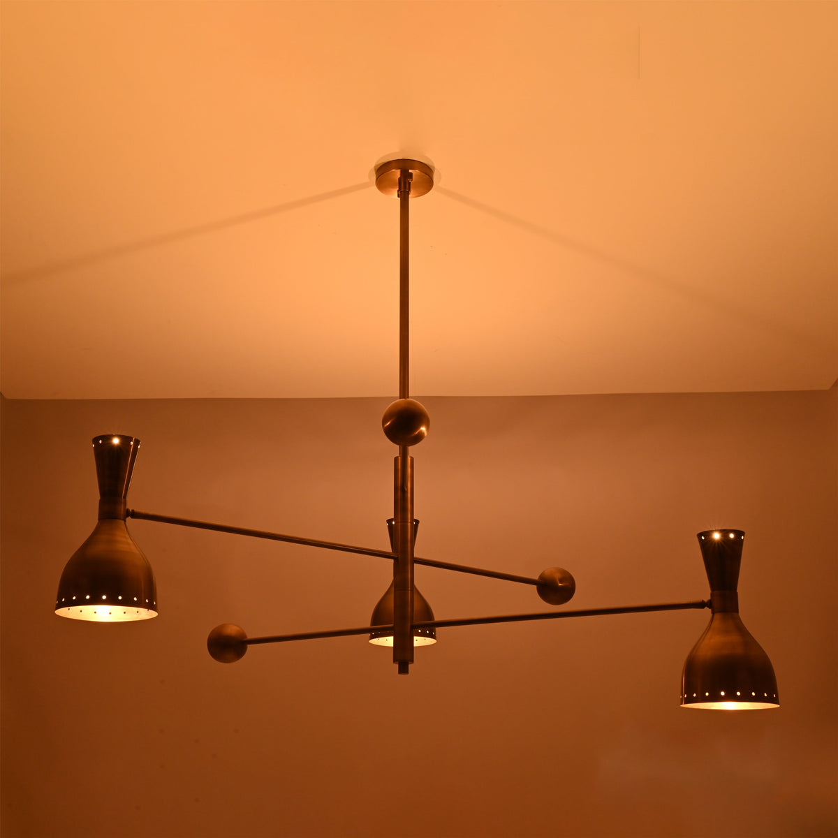 Diabolo brass chandelier illuminated in warm ambient light, creating a soft indirect glow ideal for living and dining spaces.