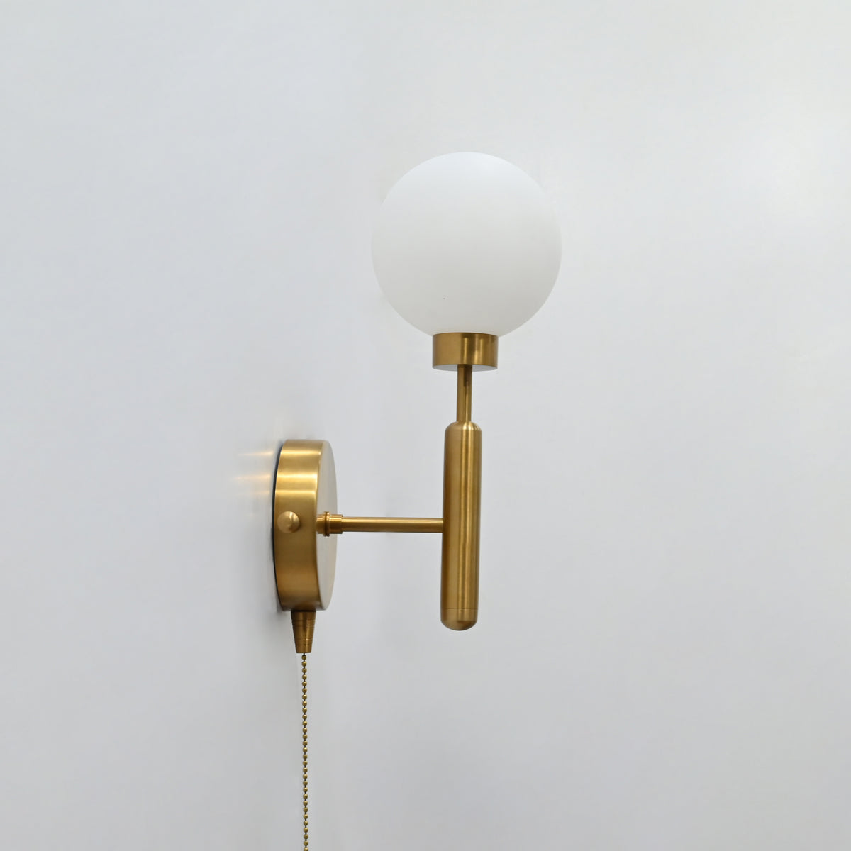 Raw brass pull-chain globe wall sconce with round frosted globe, vertical stem, and side-view mounting plate.