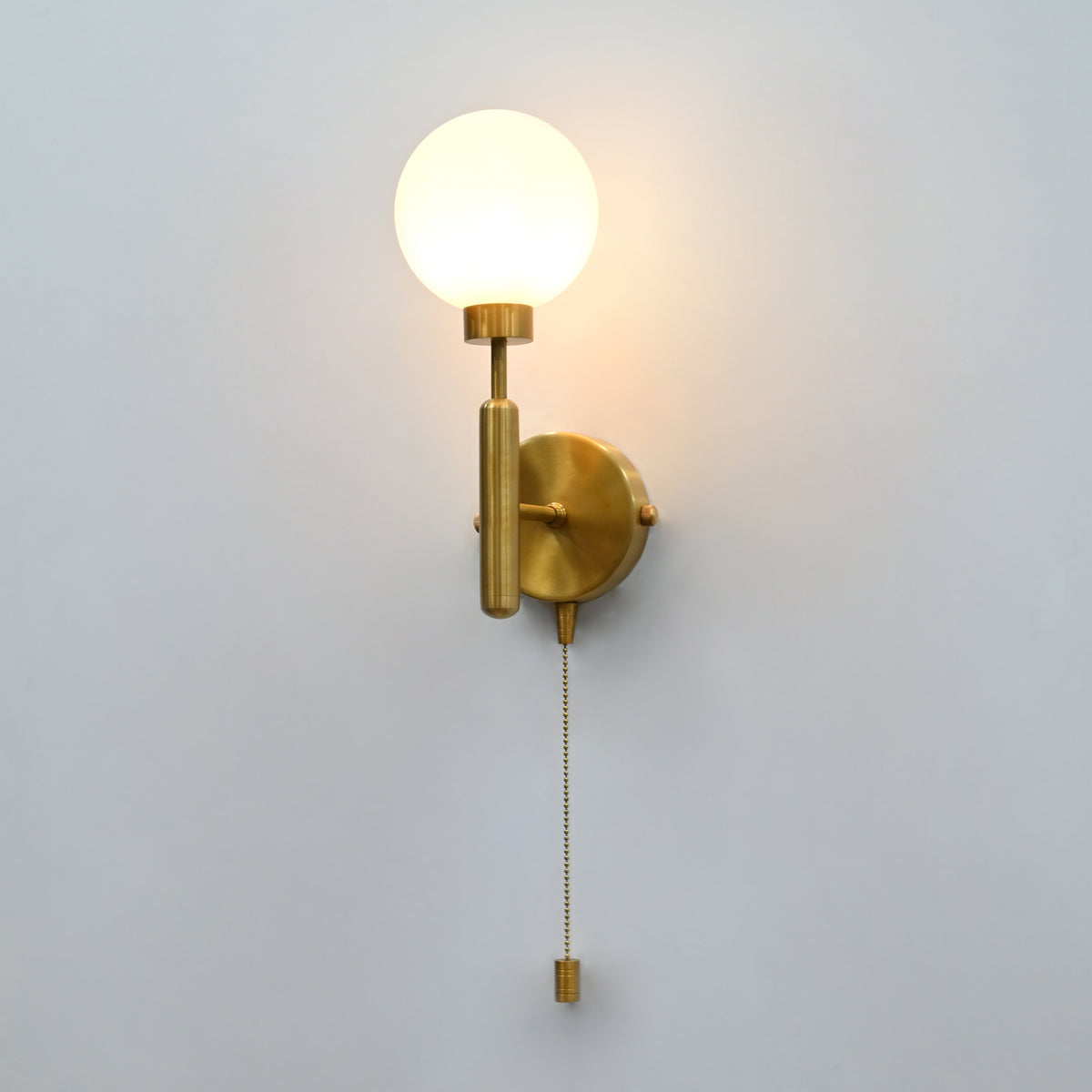 Illuminated raw brass globe wall sconce with round opal glass globe, vertical stem, round backplate and pull chain
