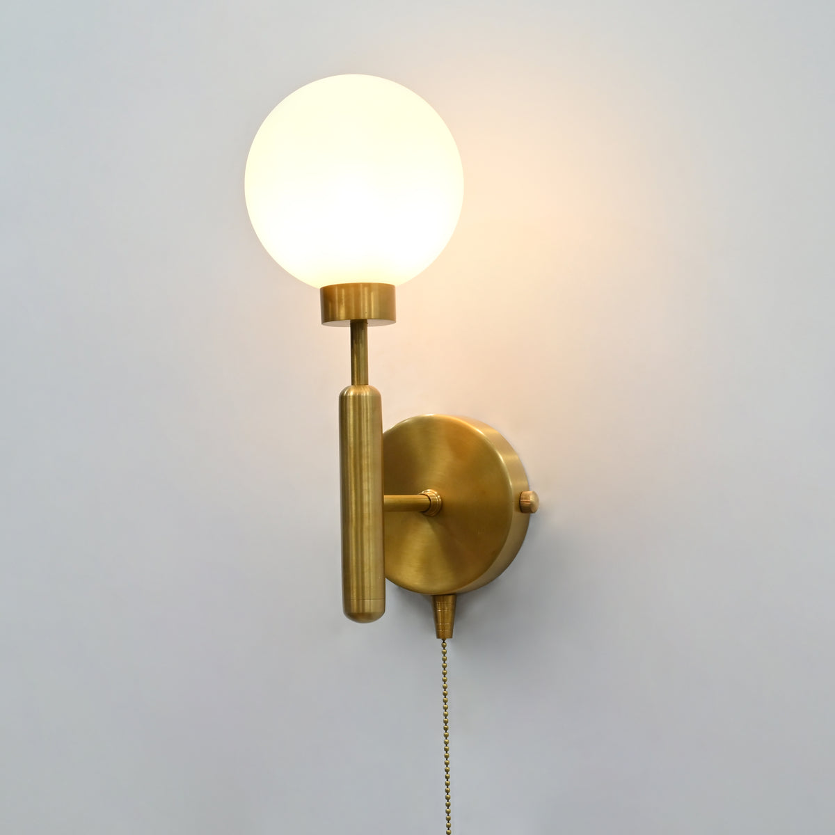 Illuminated raw brass wall sconce with round frosted globe, vertical stem, circular backplate, and pull chain.