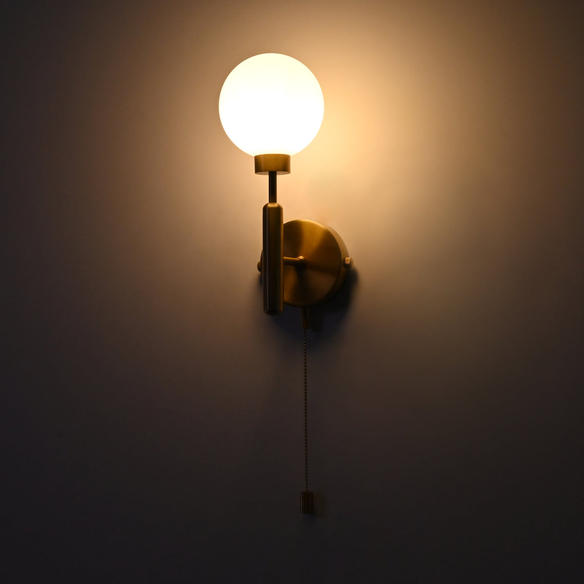 Illuminated raw brass wall sconce with round opal glass globe, vertical stem, circular backplate, and pull chain.