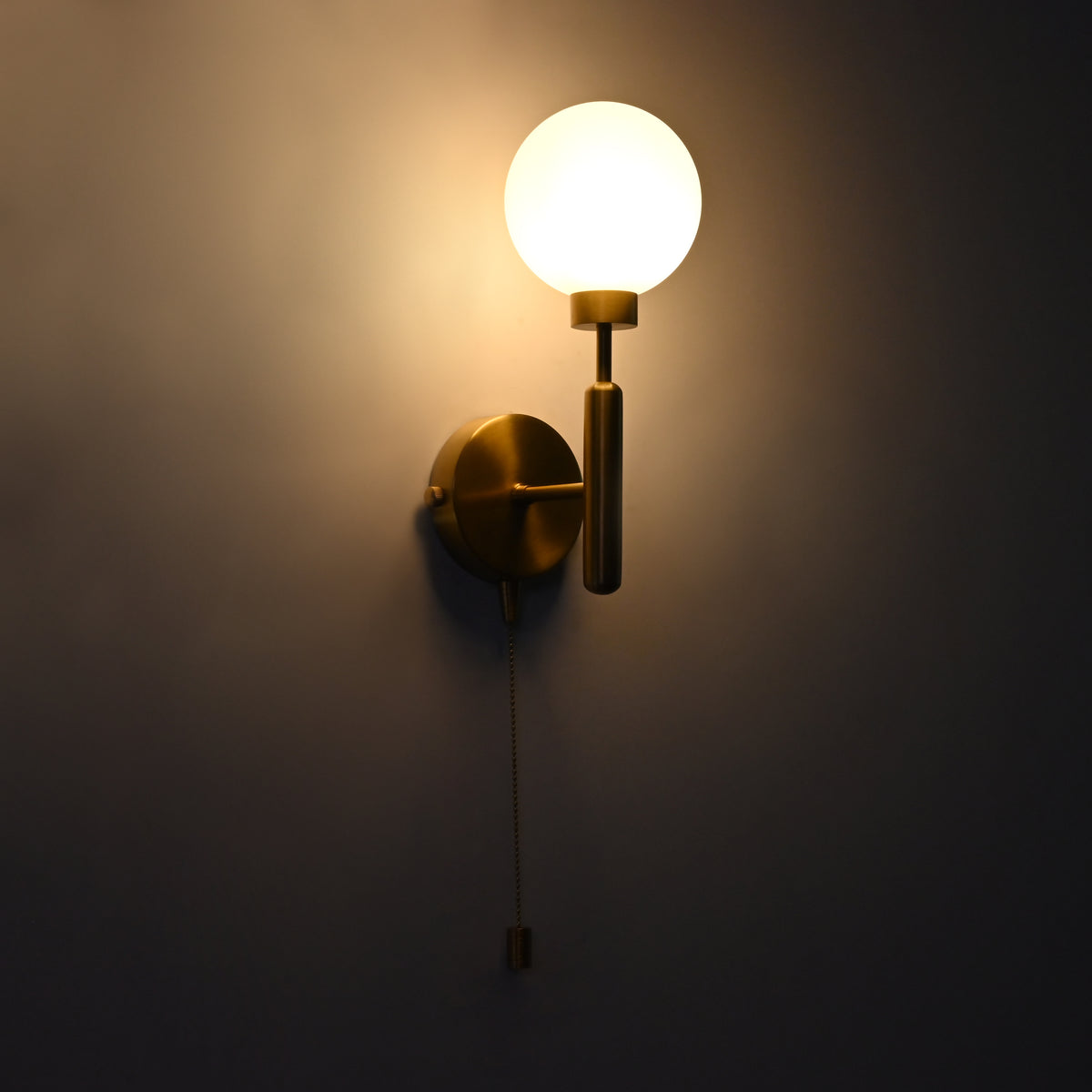 Illuminated raw brass pull chain globe wall sconce with round opal glass globe, vertical stem, and circular backplate.