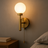 Illuminated raw brass globe wall sconce with round opal globe, vertical stem and pull chain beside a bed and wood nightstand.