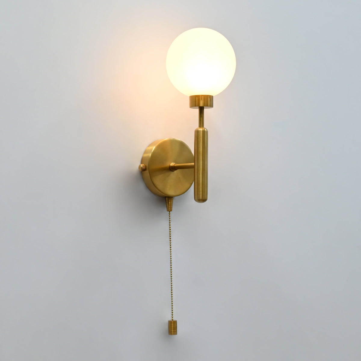 Raw brass wall sconce with round frosted glass globe shade, illuminated, and hanging pull chain switch, shown against a plain wall.